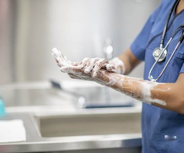 Female medical personnel scrubs hands and arms with disinfectant soap during the Covid-19 outbreak.  She is dressed in blue scrubs with a stethoscope around her neck.