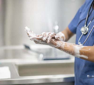 Female medical personnel scrubs hands and arms with disinfectant soap during the Covid-19 outbreak.  She is dressed in blue scrubs with a stethoscope around her neck.